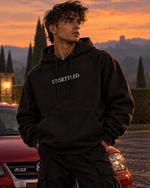Front of the black UNSETTLED Core Hoodie on a model at dusk, with a small white UNSETTLED. print on the chest